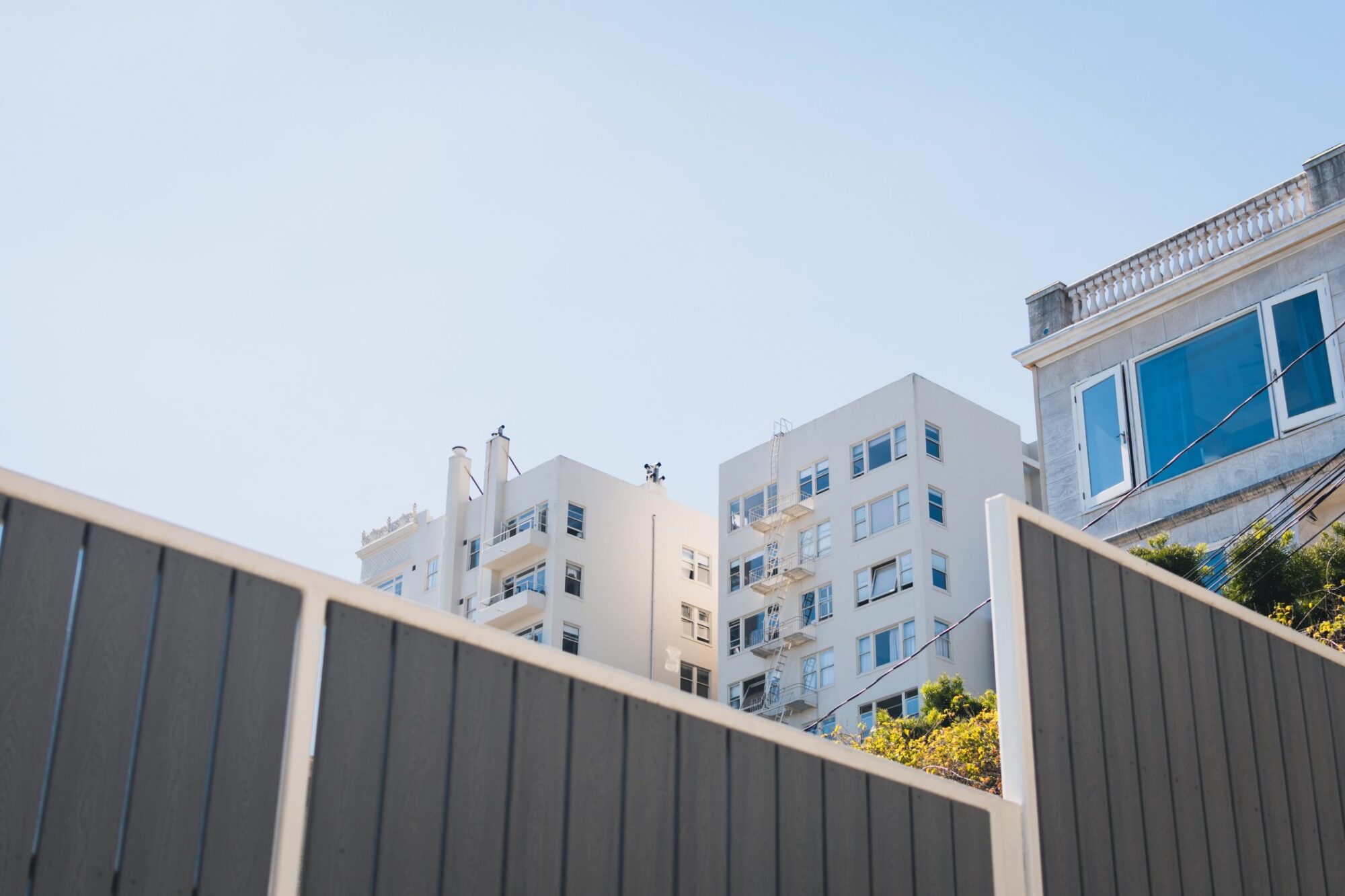 The image shows a view of urban buildings under a clear blue sky. The buildings appear to be residential apartments or similar structures. In the foreground, there's a grey fence partially obstructing the view. The architecture suggests a dense urban environment, potentially in a city. There's some foliage to the right, indicating some greenery in the area. The angle and composition of the photo create an interesting contrast between the foreground fence and the buildings in the background.