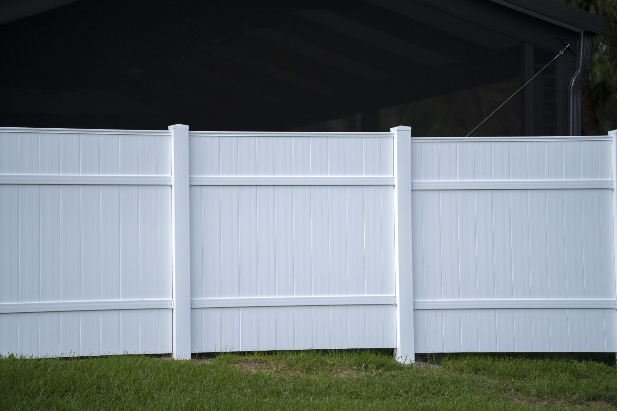 The image shows a white vinyl privacy fence with vertical panels. It's standing on a grass lawn, and behind the fence, you can see the side of a house with a dark roof. To the right side, there's a downspout attached to the house, bending towards the fence. The fence has a visible post in the centre and the overall setting appears to be a residential backyard or similar domestic outdoor area.
