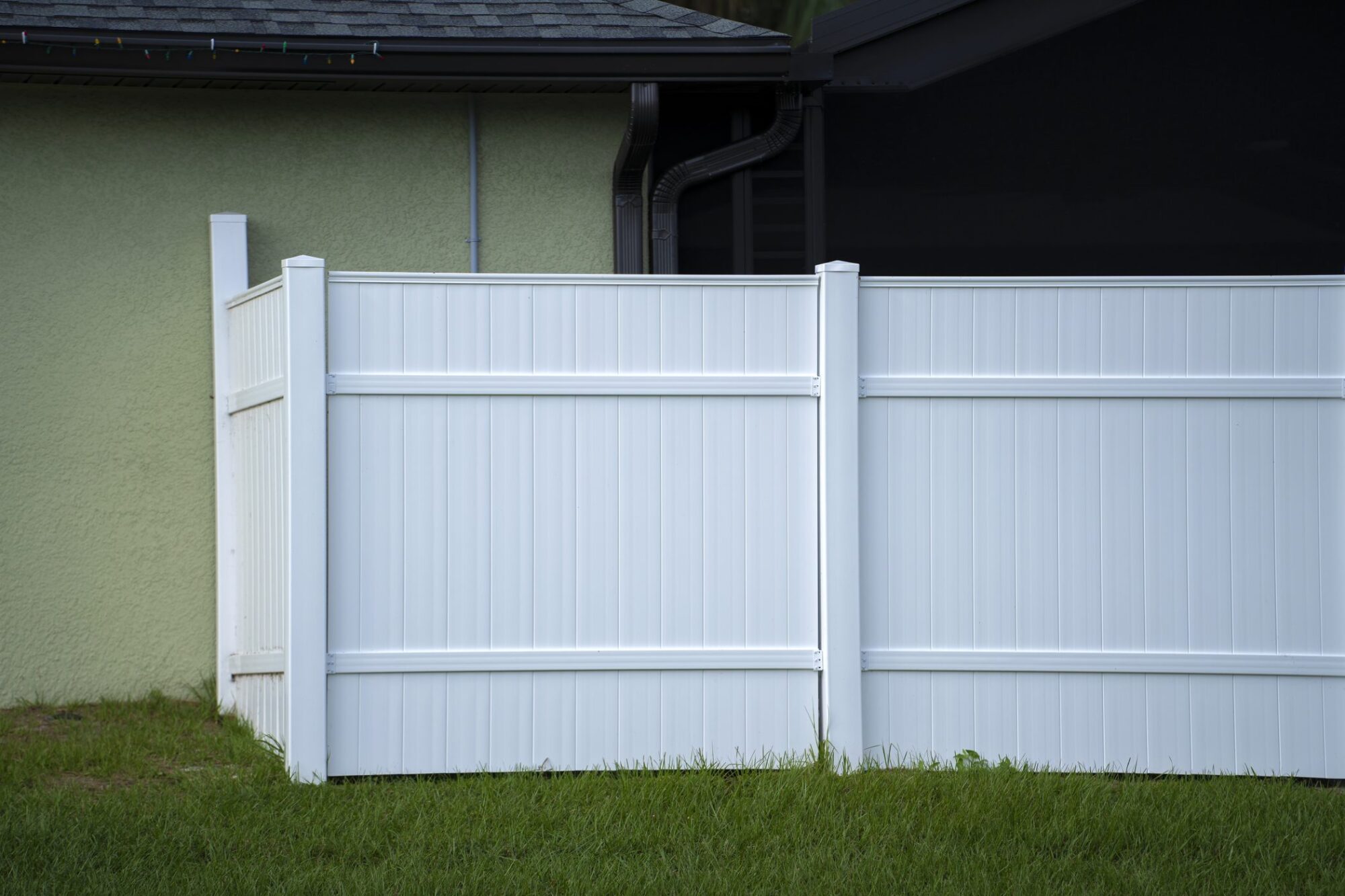 The image shows a section of a white vinyl privacy fence alongside a house. The fence is relatively tall, constructed with vertical panels, and designed to provide privacy and possibly noise reduction for the property it surrounds. In the background, there's the side of a house with light green exterior walls, a dark roof, and a downspout or gutter pipe visible. On the left edge of the image, you can see a part of what seems to be either a gate or a fence post. The fence is set on green grass, indicating an outdoor residential setting.