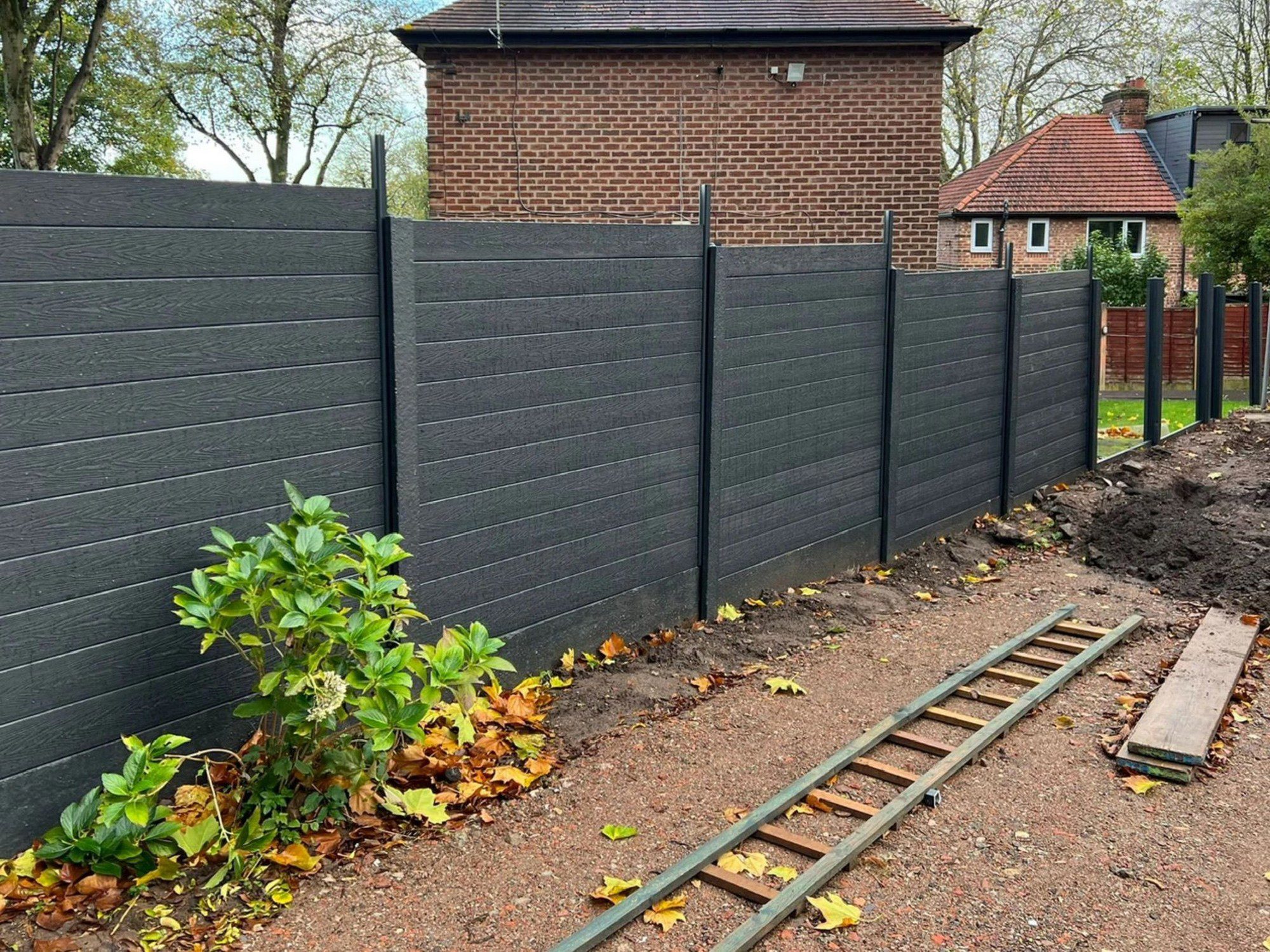 The image shows a section of a garden or outdoor area. In the forefront, there's a dark gray or black composite fence that appears to be new and extends throughout the image. To the left, there's a green shrub with some scattered fallen leaves around its base, indicating it might be autumn. There's also a section of soil that looks freshly dug or disturbed along the fence line, possibly where the fence was recently installed or for another gardening purpose. On the ground are two parallel metal tracks or beams, in front of which lies a single, long concrete slab. In the background, there's a brown brick house with a chimney, and another house visible behind the fence. The sky is overcast, suggesting it may be a cloudy day.