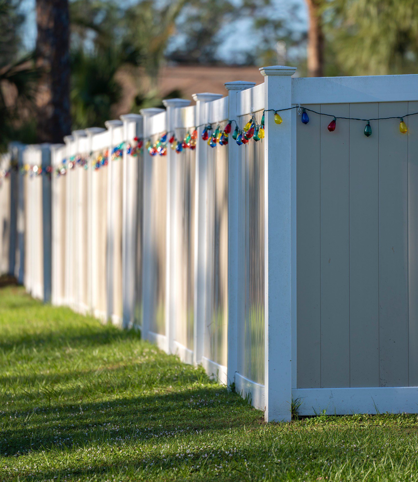 The image shows a long white fence stretching across the frame with a string of colorful lights draped along the top. These lights appear to be outdoor decorative lights, commonly used during holiday seasons or for festive occasions. The setting seems to be a well-maintained lawn or garden, indicated by the neatly cut grass and a few small plants at the base of the fence. In the background, there's some greenery and what looks like a clear, sunny sky.