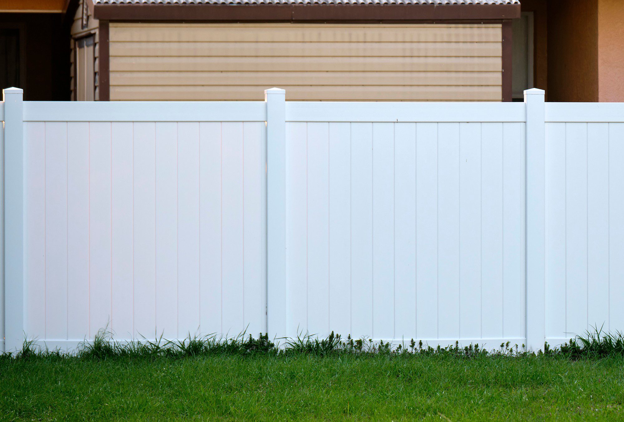 The image shows a section of a white vinyl fence installed on a well-maintained grass lawn. The fence has vertical panels, capped with pointed post tops. In the background, part of a house or building can be seen, with a brown roof and a partially visible window covered with closed beige-coloured blinds. The overall scene suggests a suburban residential area. There are no people in the image.